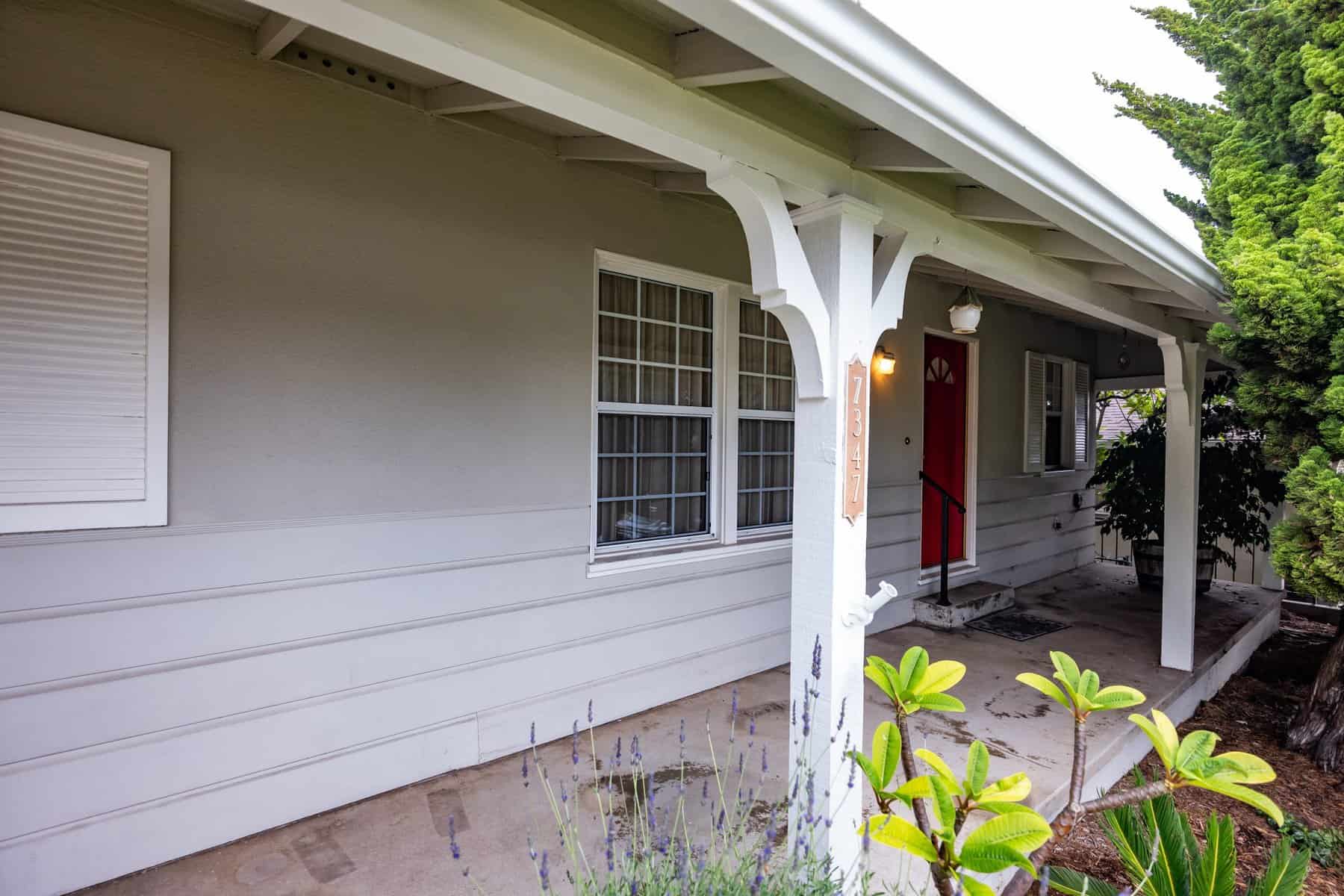 Front porch of a gray house with a red door, white trim, and surrounding greenery